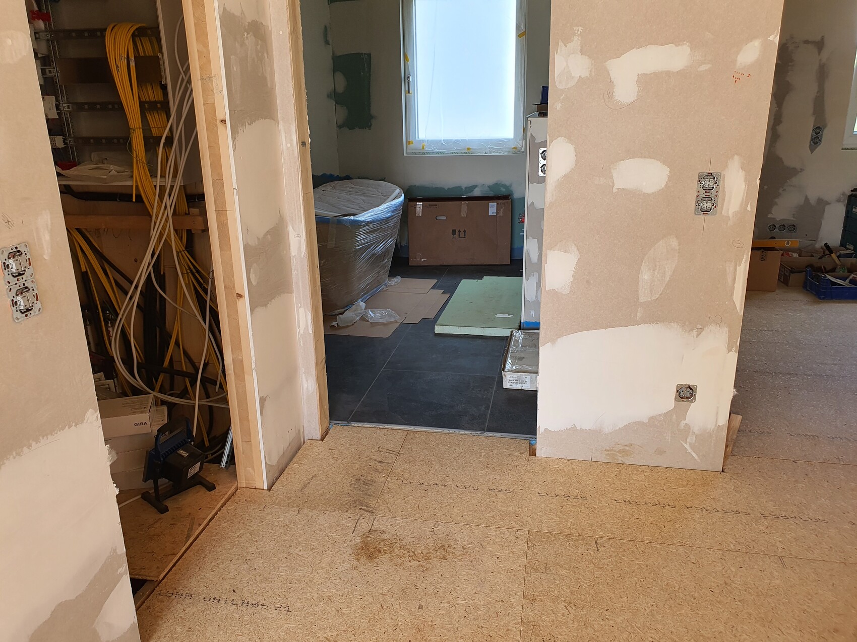 Baustelle: links Kabelbündel, rechts verputzte Wände, verpackte Gegenstände im Raum.