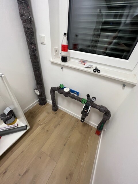 Insulated pipes with green valve handles under window, wooden floor.