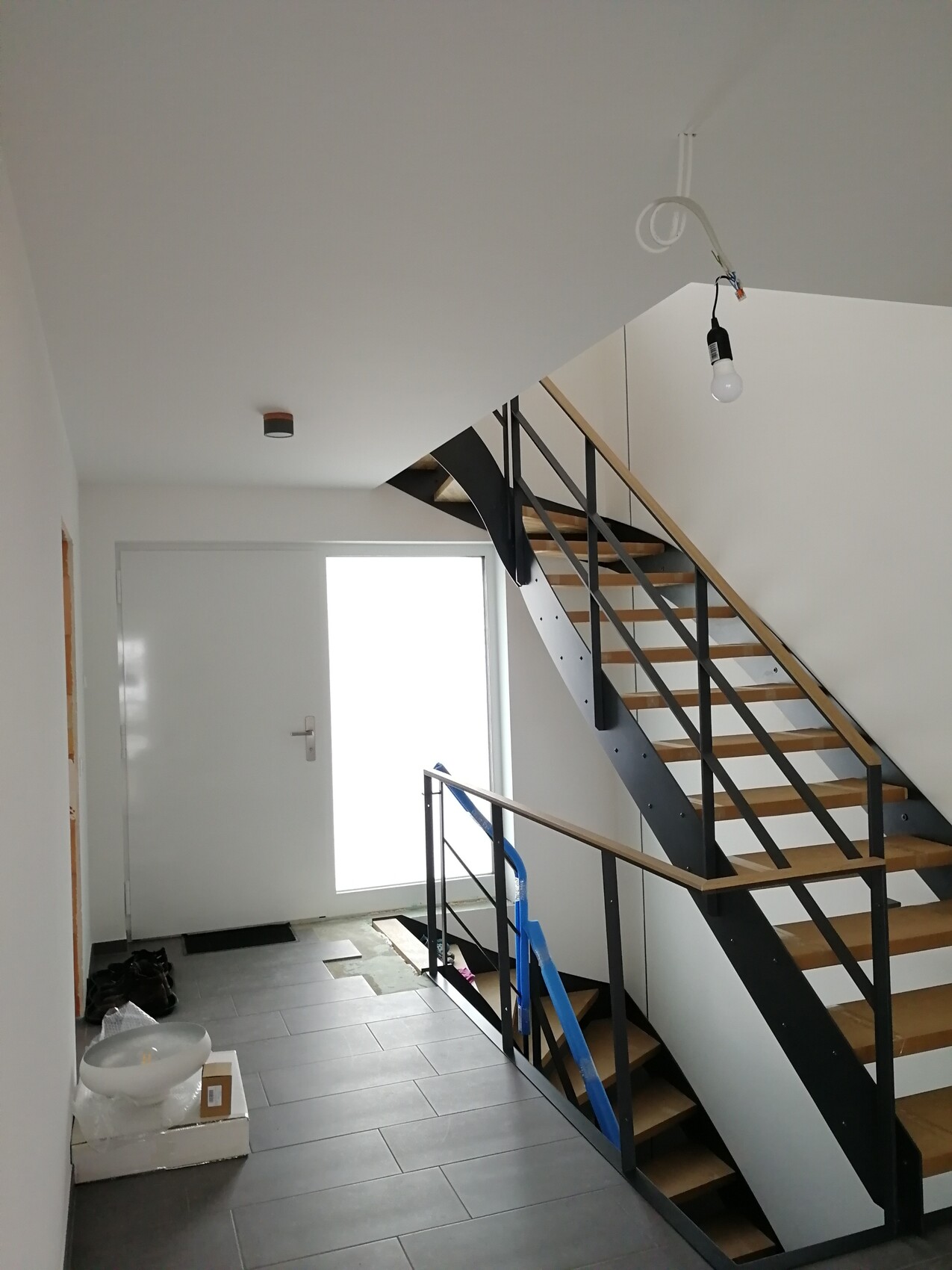 Bright entrance area with open wooden staircase and black metal frame; construction materials on the floor.