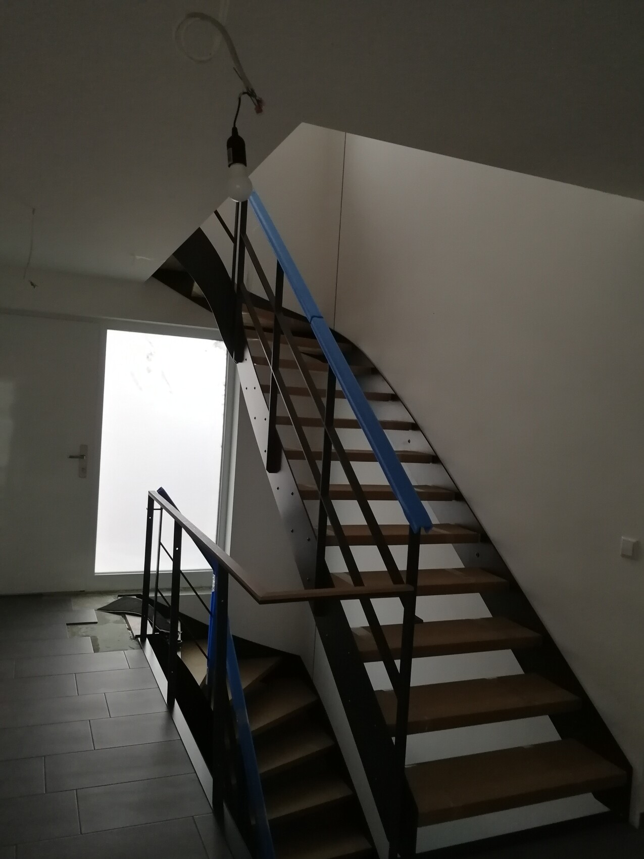 Curved metal staircase with wooden steps, blue protective film on the railing, construction lighting.