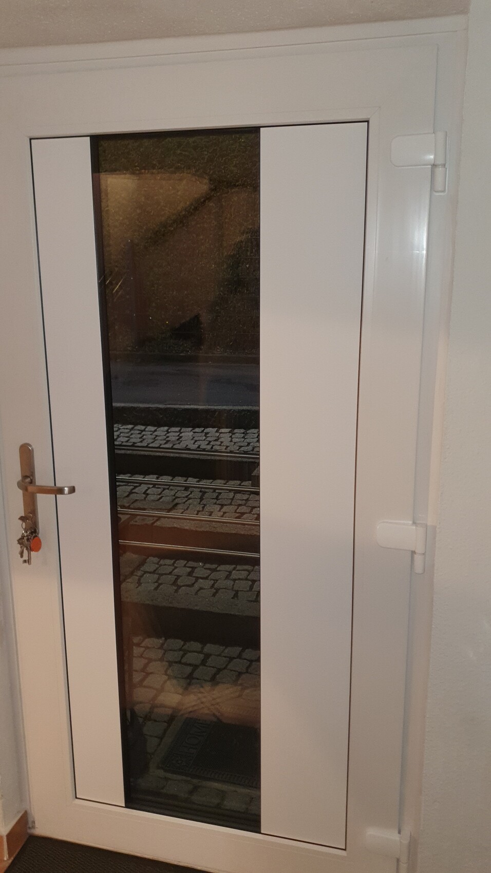 White door with narrow vertical glass window, handle with key, view outside onto pavement.