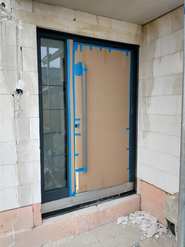 Door with glass front on construction wall, cardboard covering and blue painter’s tape on the frame.