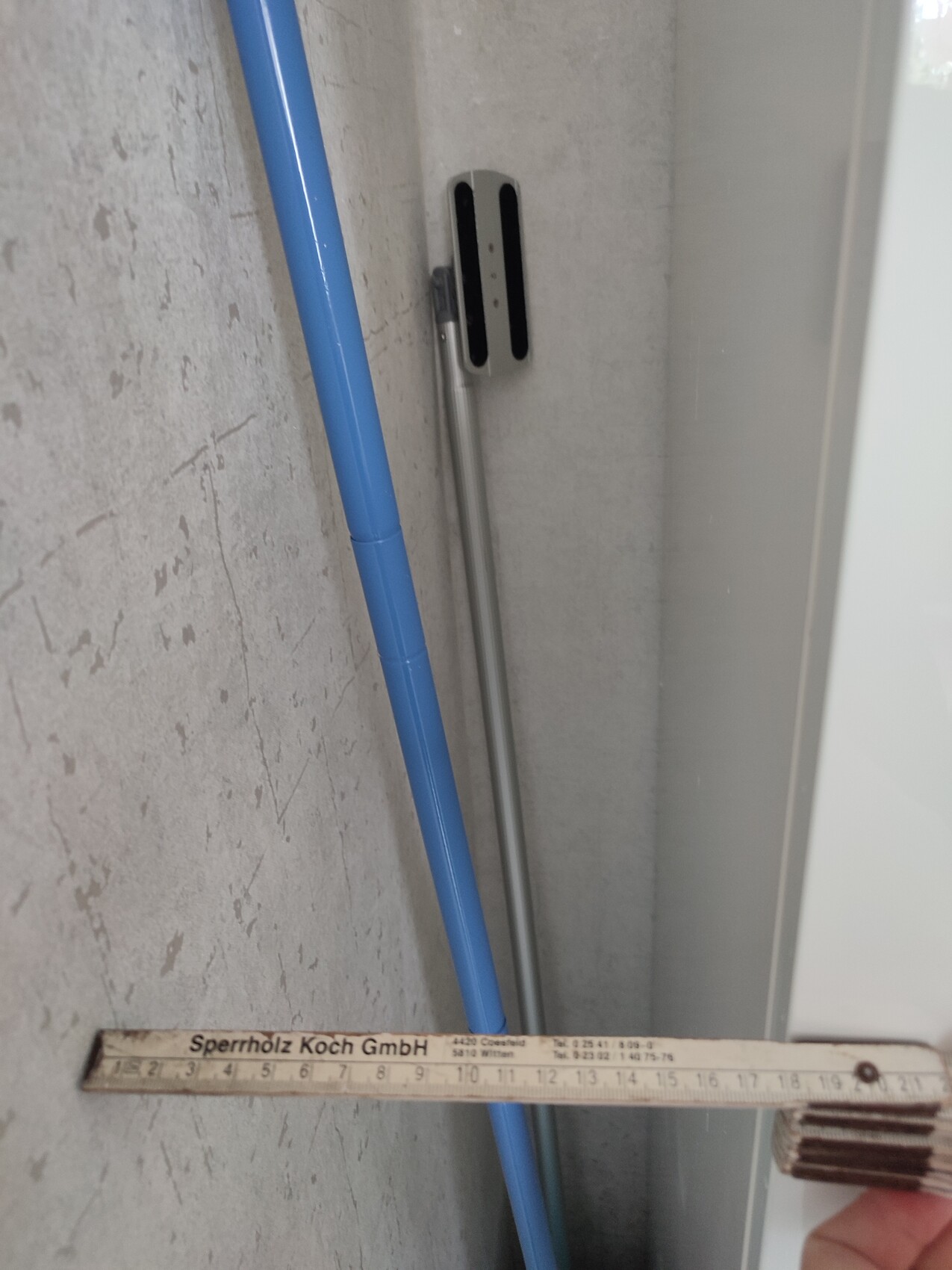 Blue cleaning stick leaning against a grey wall; a wooden folding ruler lies in the foreground.