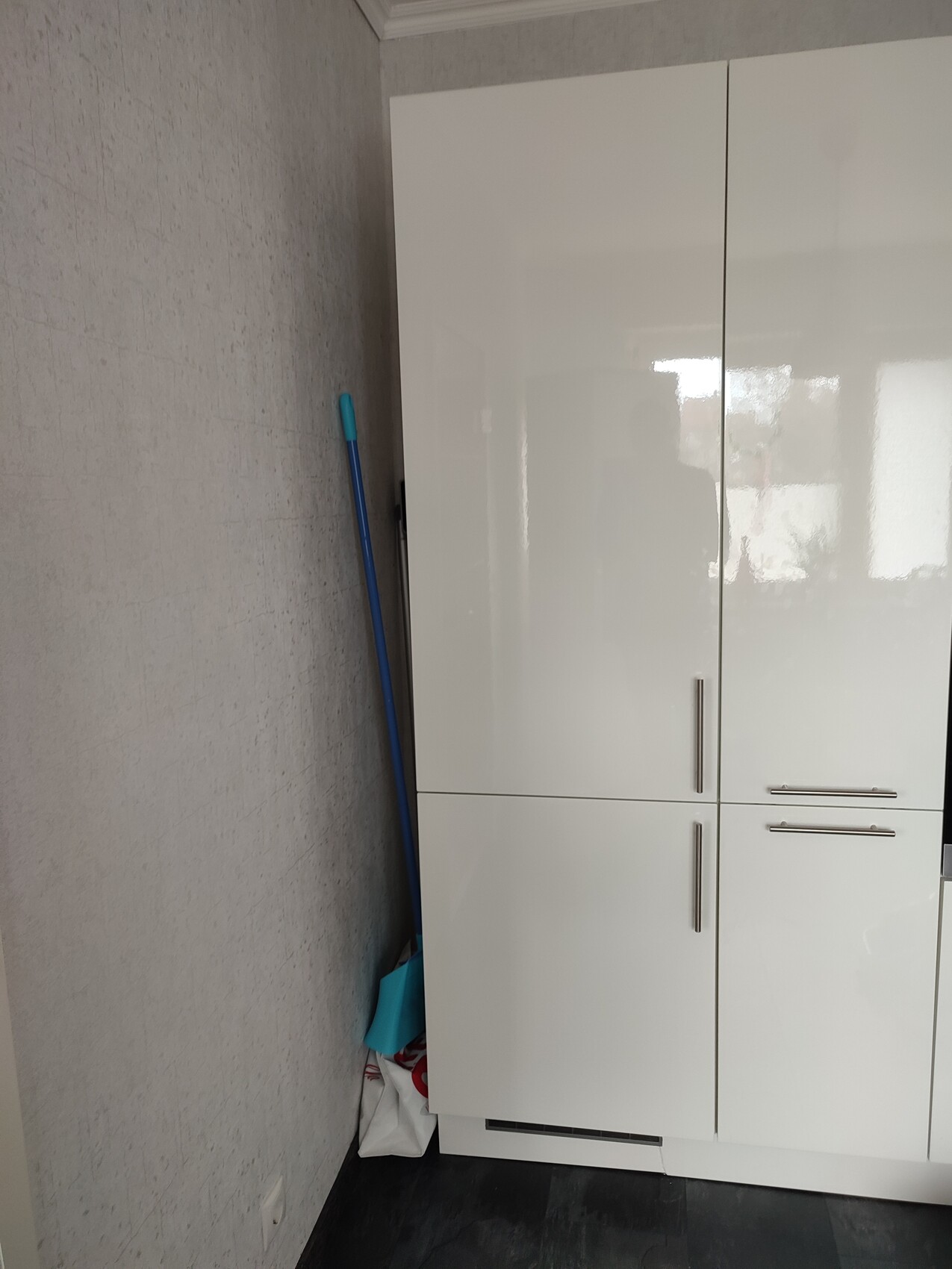 White cabinets in storage room; blue cleaning stick leaning against a grey wall, rubbish bags on the floor.