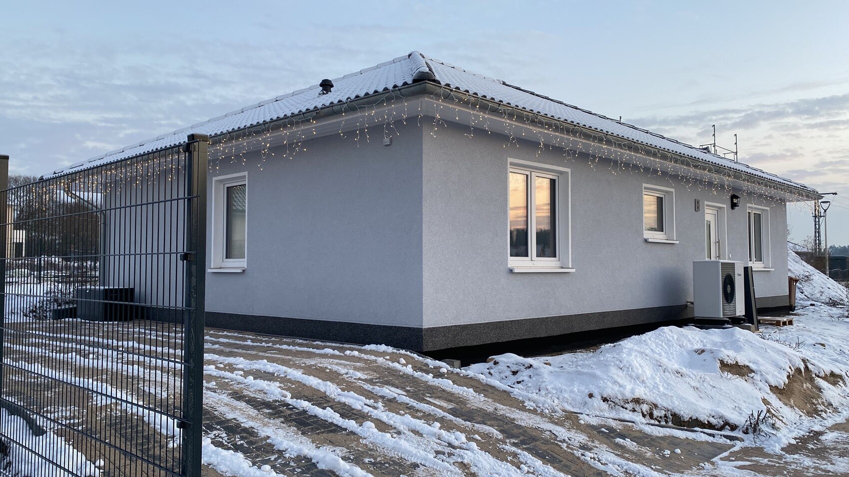 Einfamilienhaus mit grauer Fassade, Schnee, Lichterketten am Dach, Zaun im Vordergrund