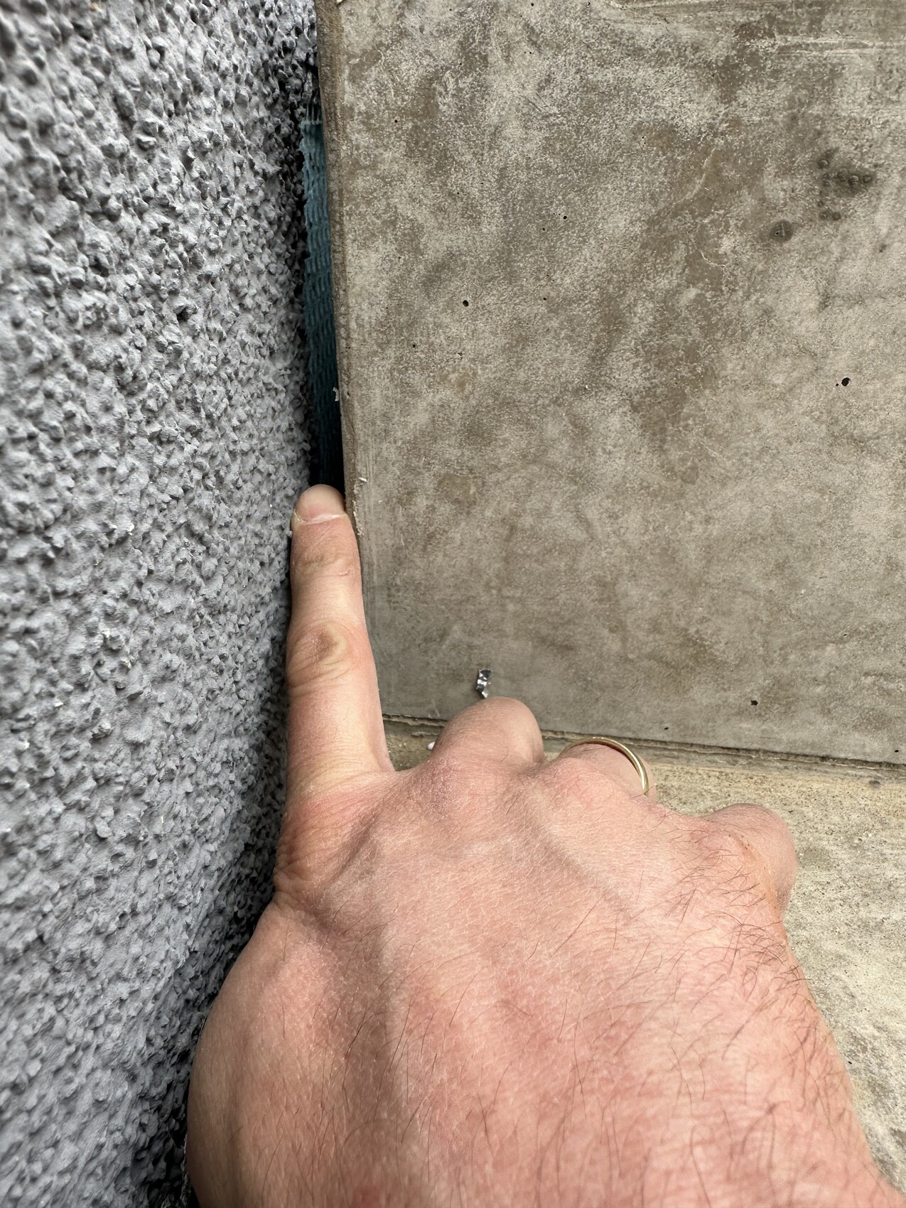 Hand with ring pointing to a small screw in the gap between rough wall and concrete slab.
