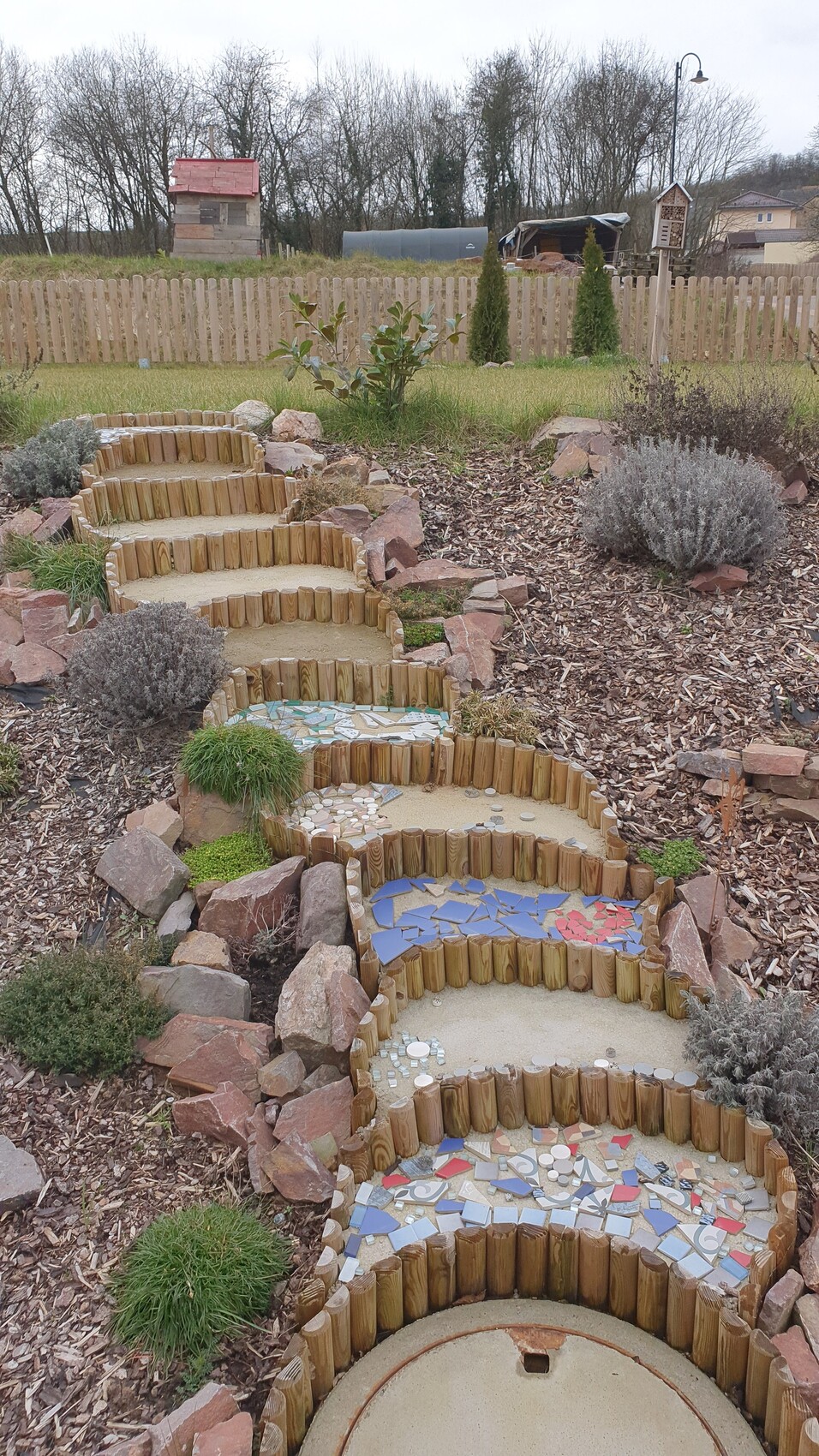 Gartenpfad mit aufeinander gestaffelten Holzstufen und bunten Fliesen-Mosaik, von Steinen umgeben.