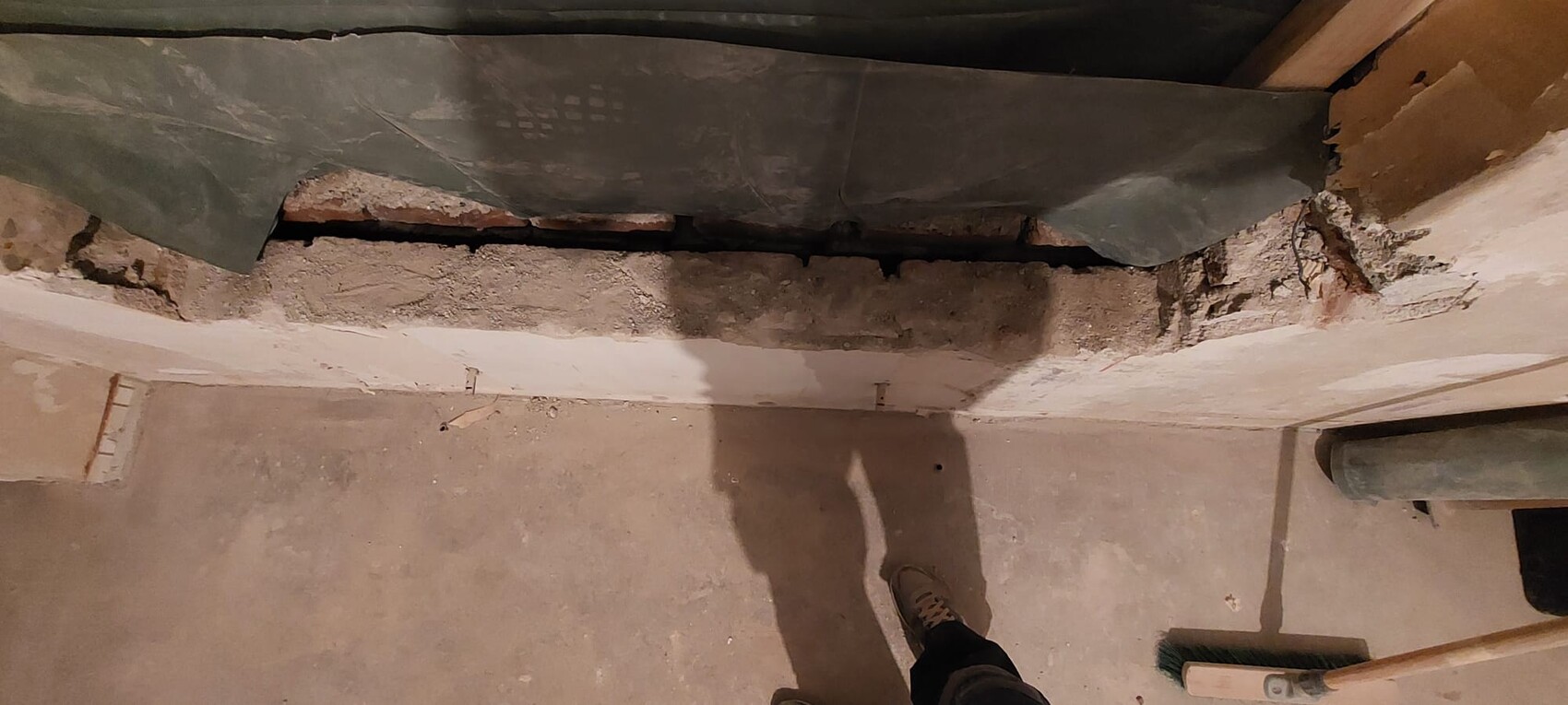 Basement construction site: damaged wall with gap, plaster flaking off; broom and shoe visible.