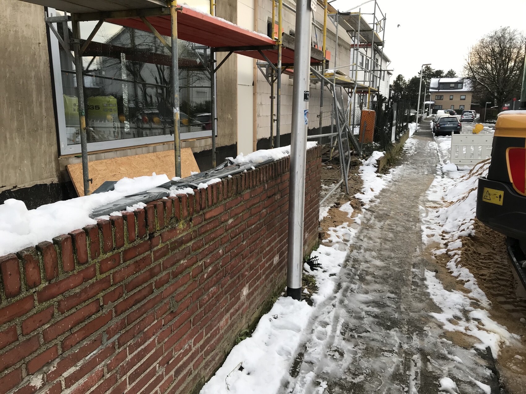 Construction site on sidewalk in winter: scaffolding, brick wall, snow, mud; yellow excavator on the right.
