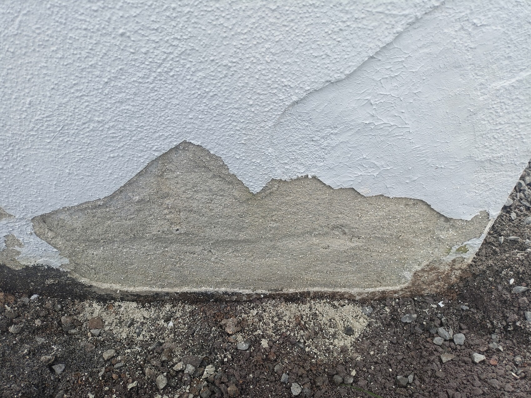 Wall with peeling plaster; grey concrete underneath, floor made of gravel and rubble.