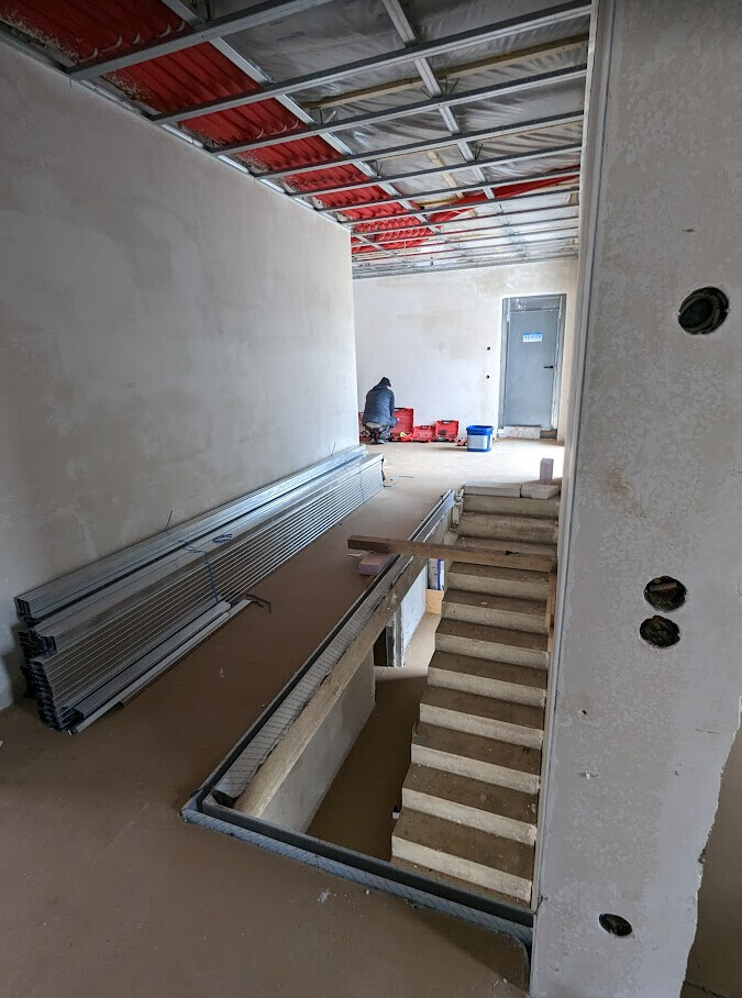 Interior construction: ceiling with metal frame, open staircase, worker by red boxes in front of door.