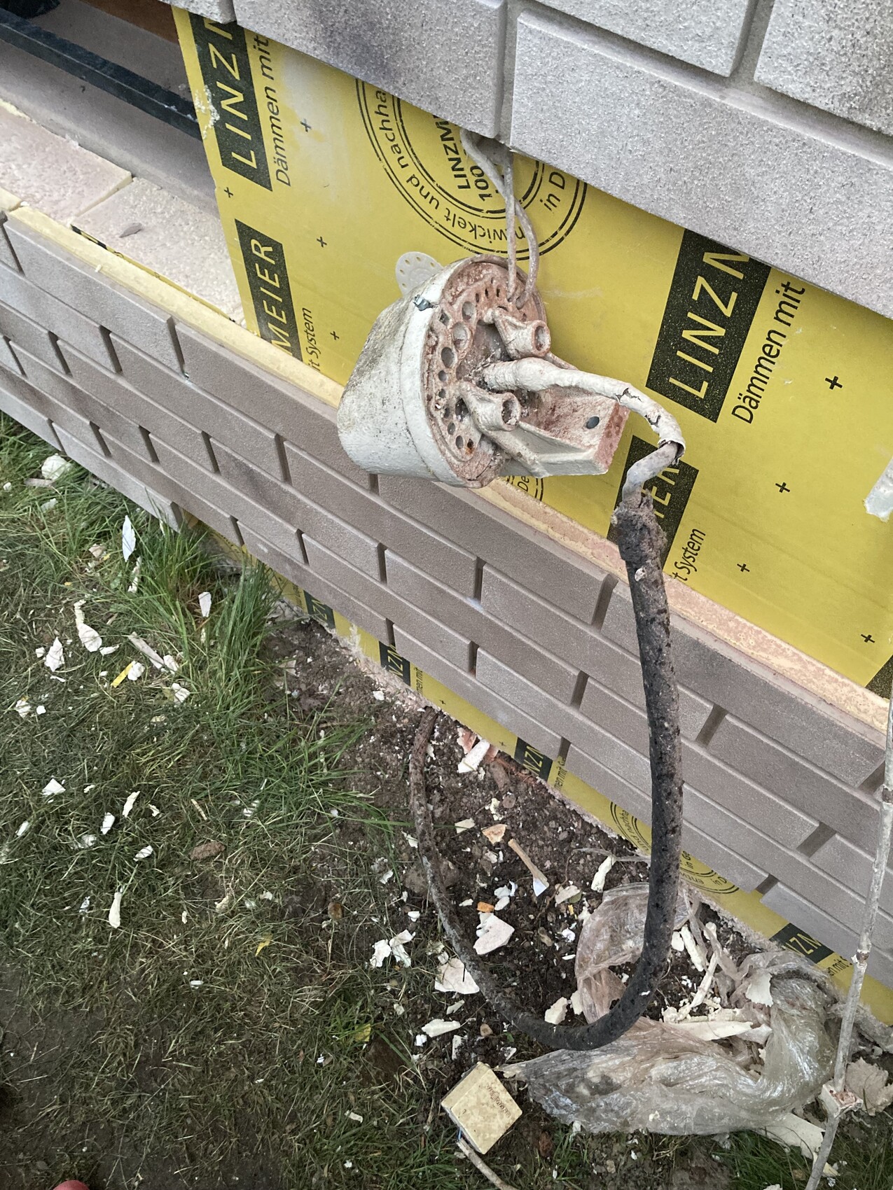 Rusty metal part with perforated disc hanging on wall; cables and construction debris lying nearby.