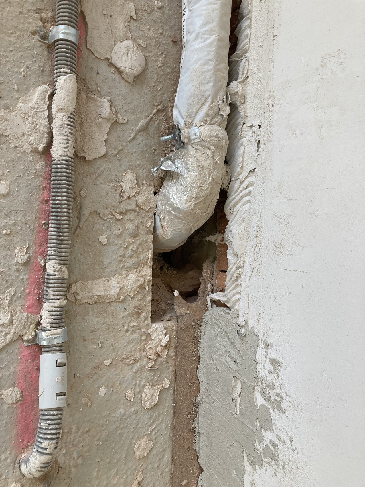 Open wall opening with insulated pipe, flexible conduit, and crumbling plaster.