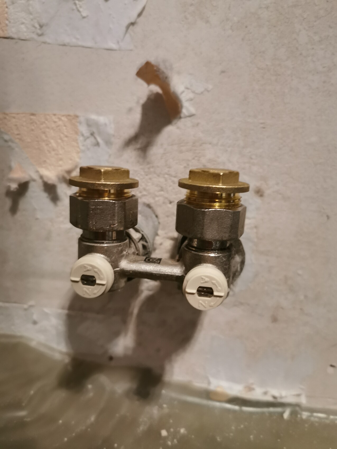 Double shut-off valve made of metal with two white rotary knobs on a gray wall.