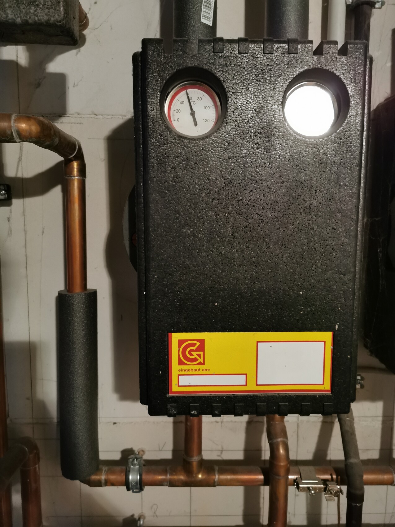 Black heating boiler on the wall with temperature display, copper pipes and yellow logo at bottom.