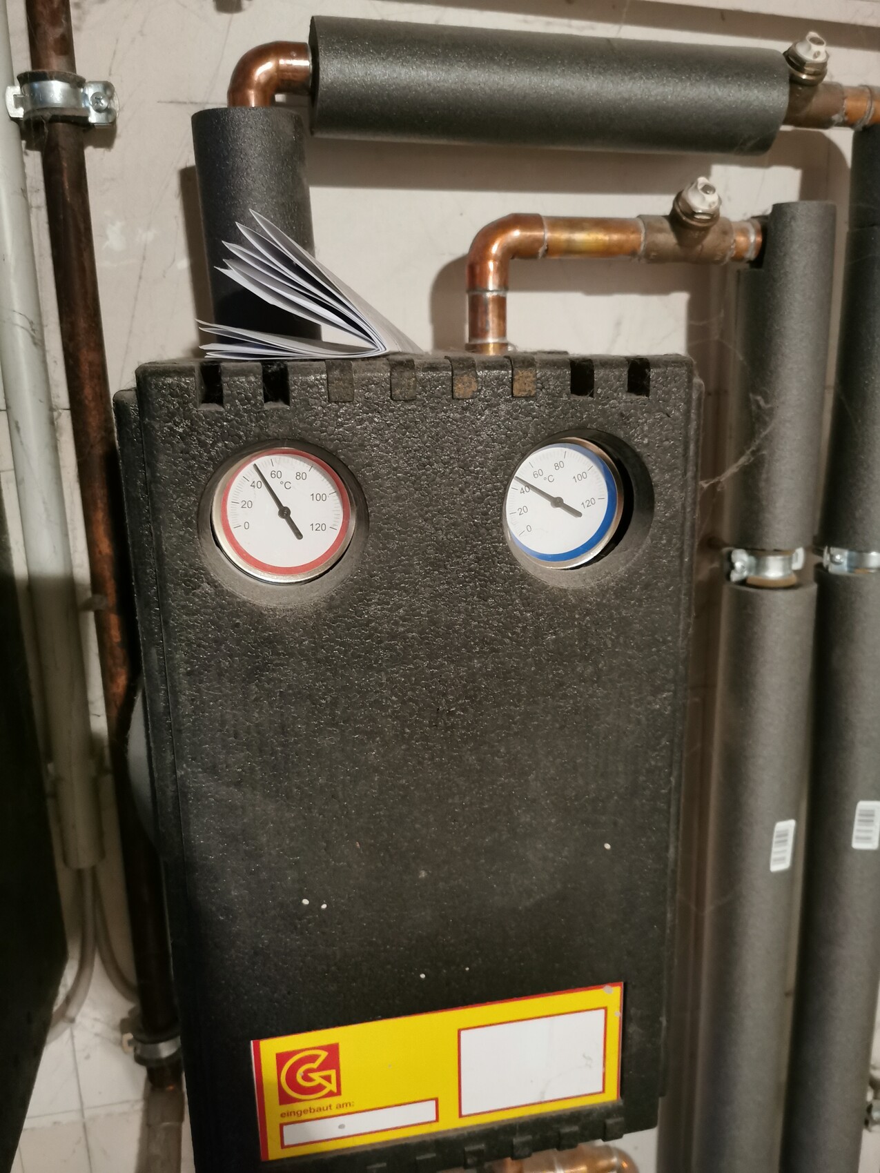 Heating boiler with two round instruments (temperature/pressure) and stack of papers on it, copper pipes beside.
