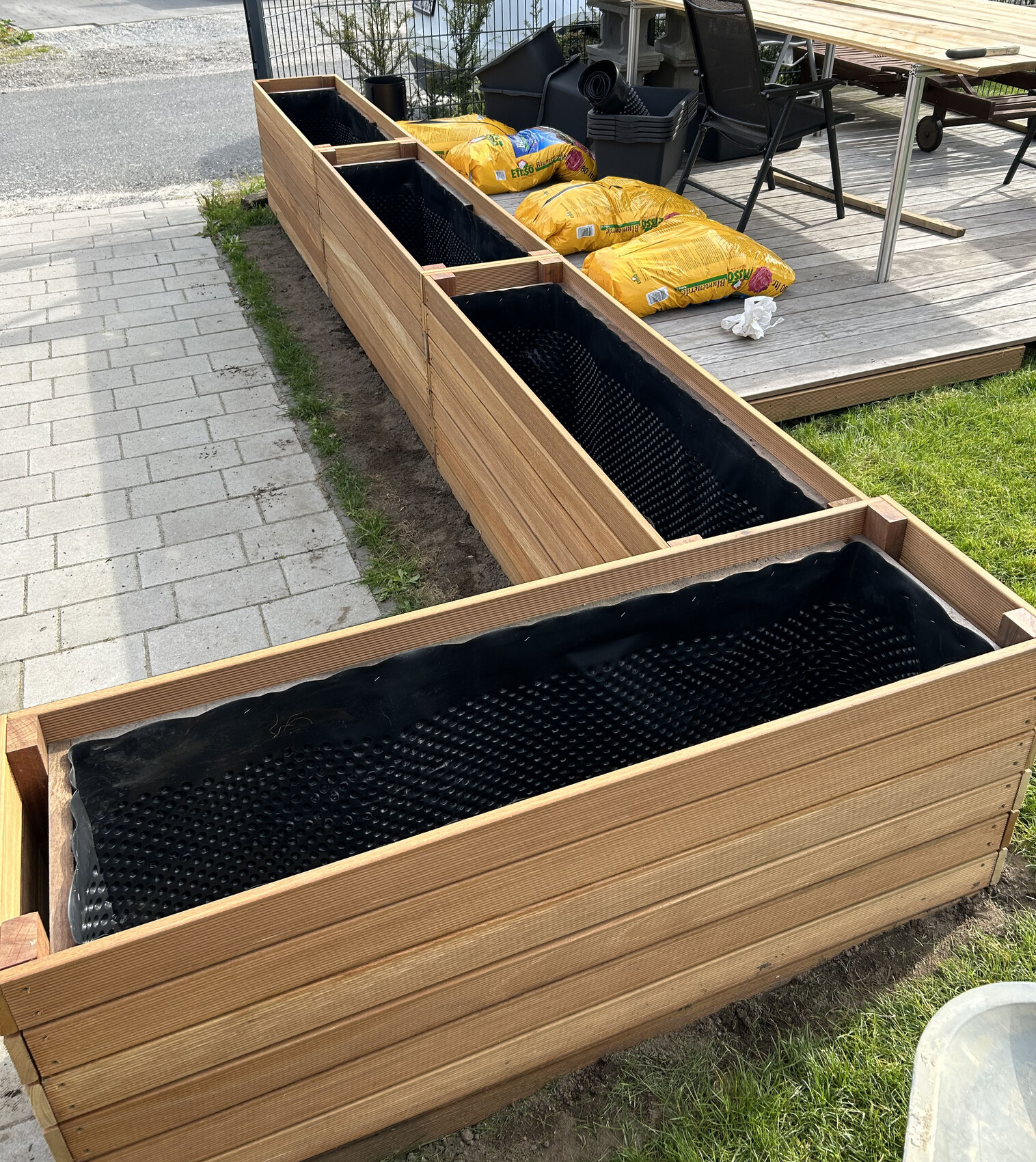 Mehrere Holzhochbeete mit schwarzer Innenfolie, neben gelben Gartensäcken auf der Holzterrasse.