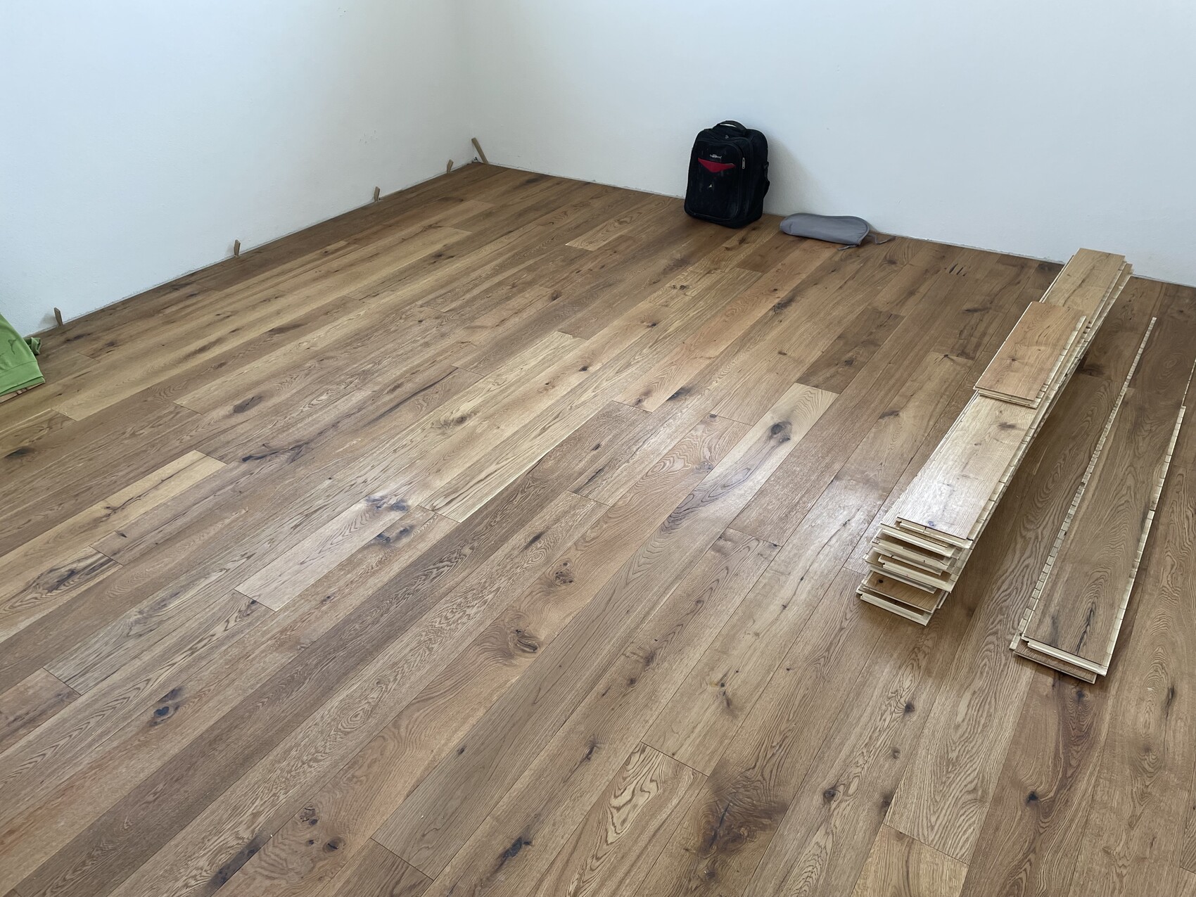 Newly installed wood floor, with a stack of planks on the right and a black backpack in the corner.