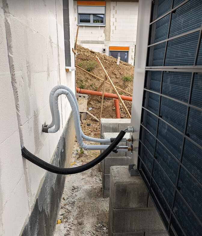 Exterior view of a construction site: pipes and cables run along a wall; earthworks in the background.