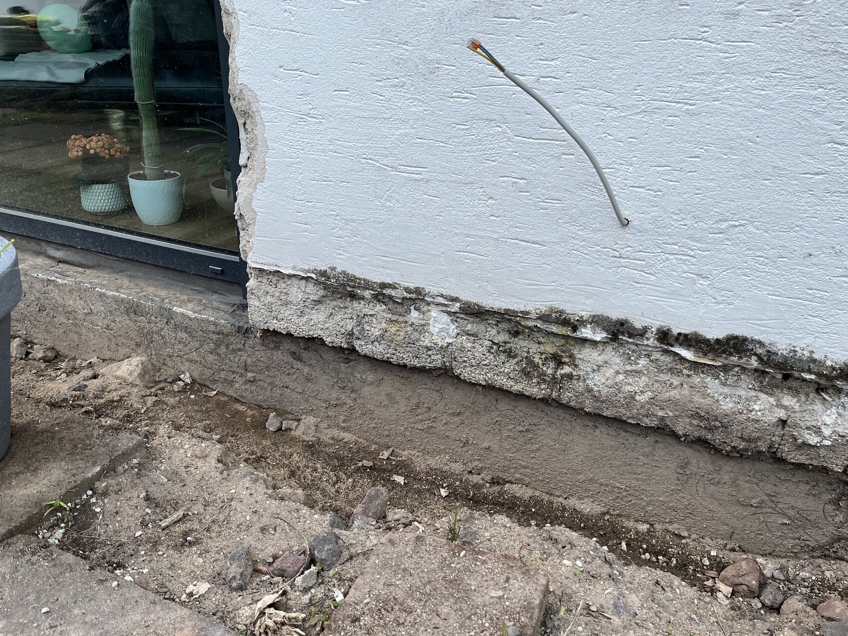 Exterior wall with exposed foundation, soil underneath, cable coming out of the wall, glass door on the left.