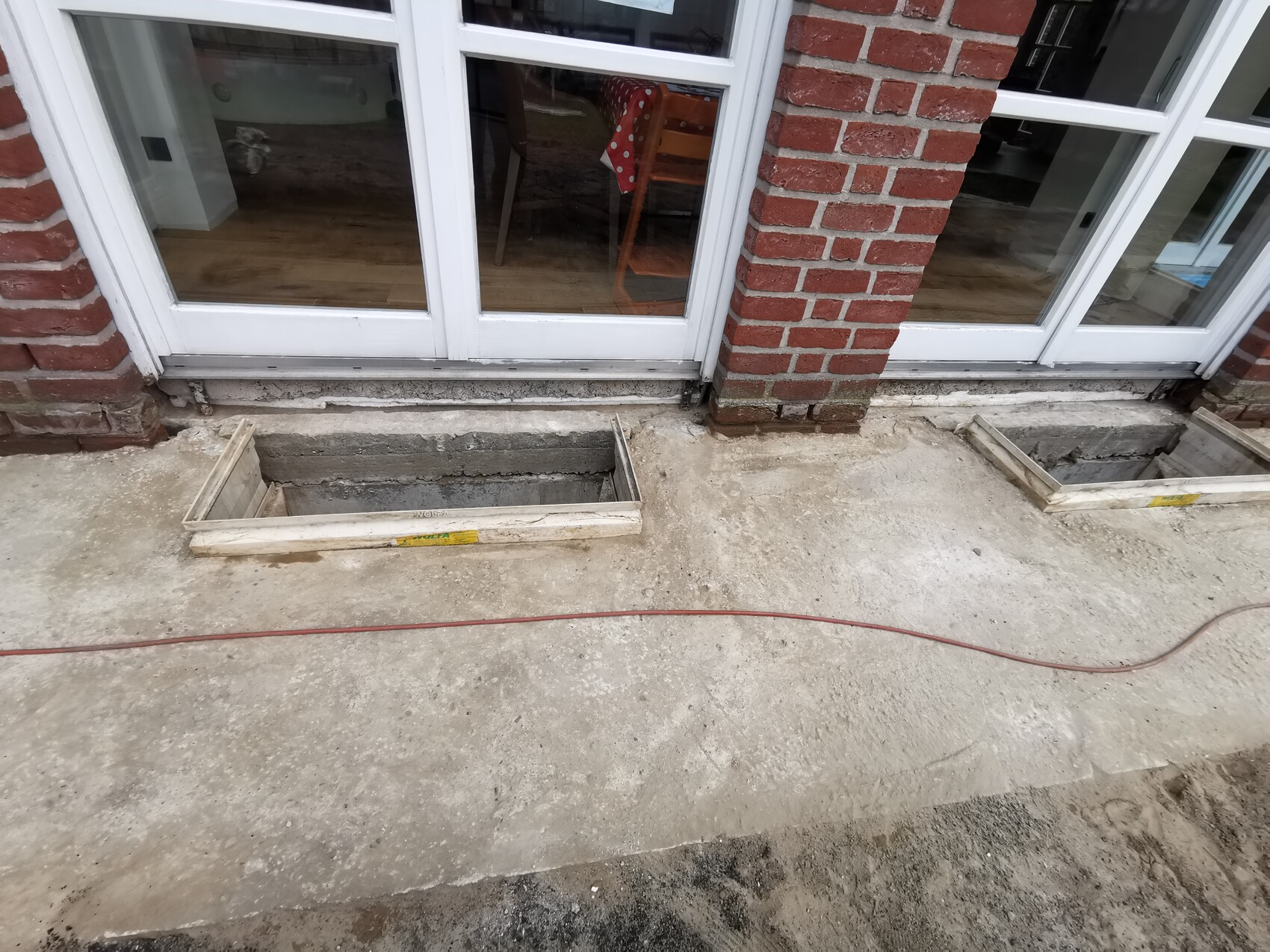 Construction site in front of glass doors: rectangular concrete pits with wooden frames, cables on the ground.