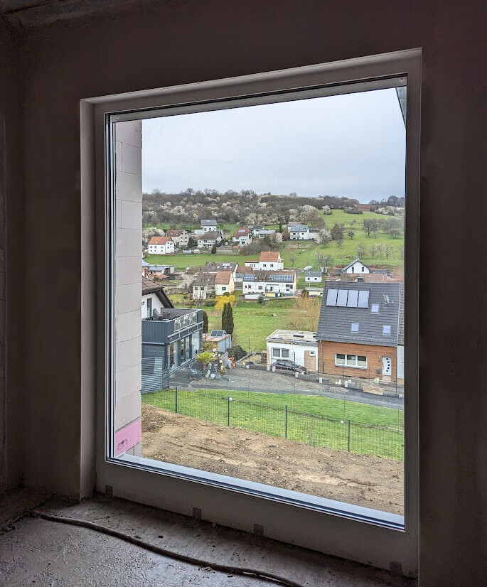 View from unfinished interior through a large window onto a suburb with houses, fields, and hills.