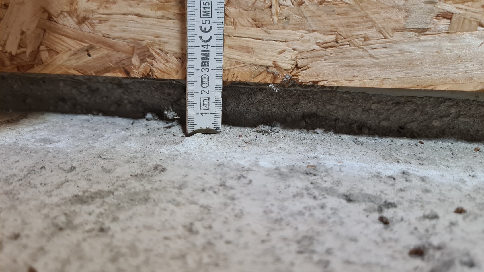 Tape-Maß steht senkrecht vor OSB-Wand über Betonboden; Staub am Boden sichtbar.