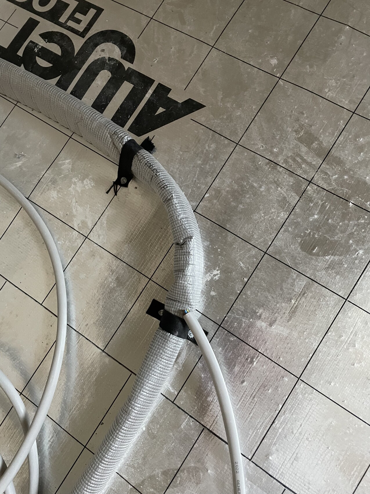 Silver flexible metal cable/duct, bent, attached to a bracket on a dusty tile floor.