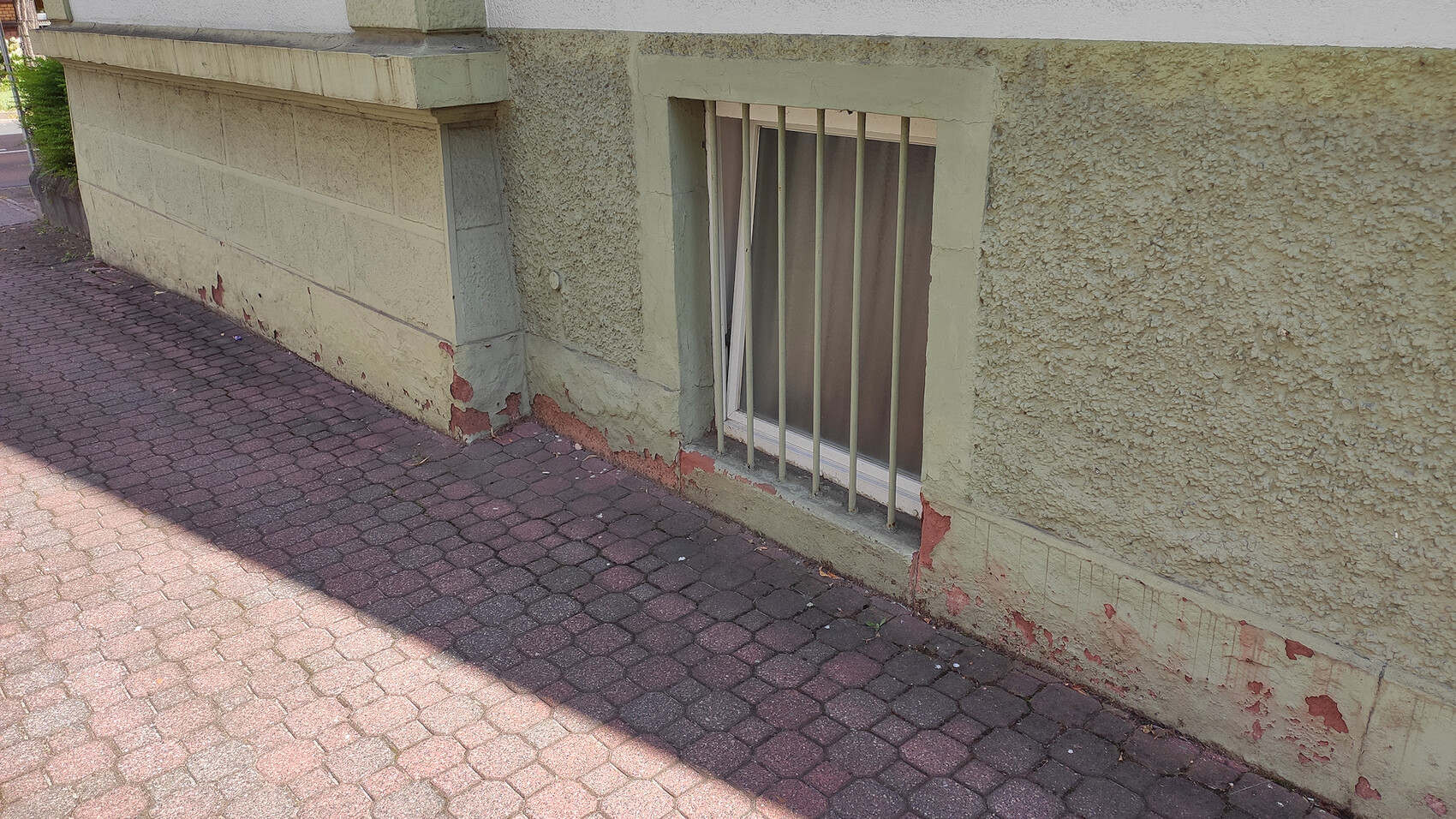Exterior wall of a building with basement window behind metal bars, green plaster, and abrasion at the base.