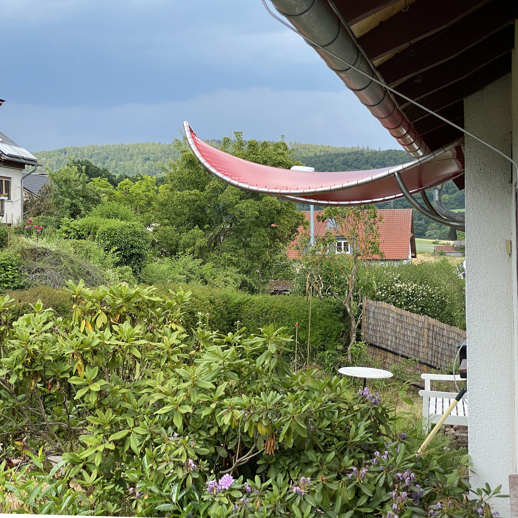 Garden with dense greenery, red curved overhang on the house, and hill in the background