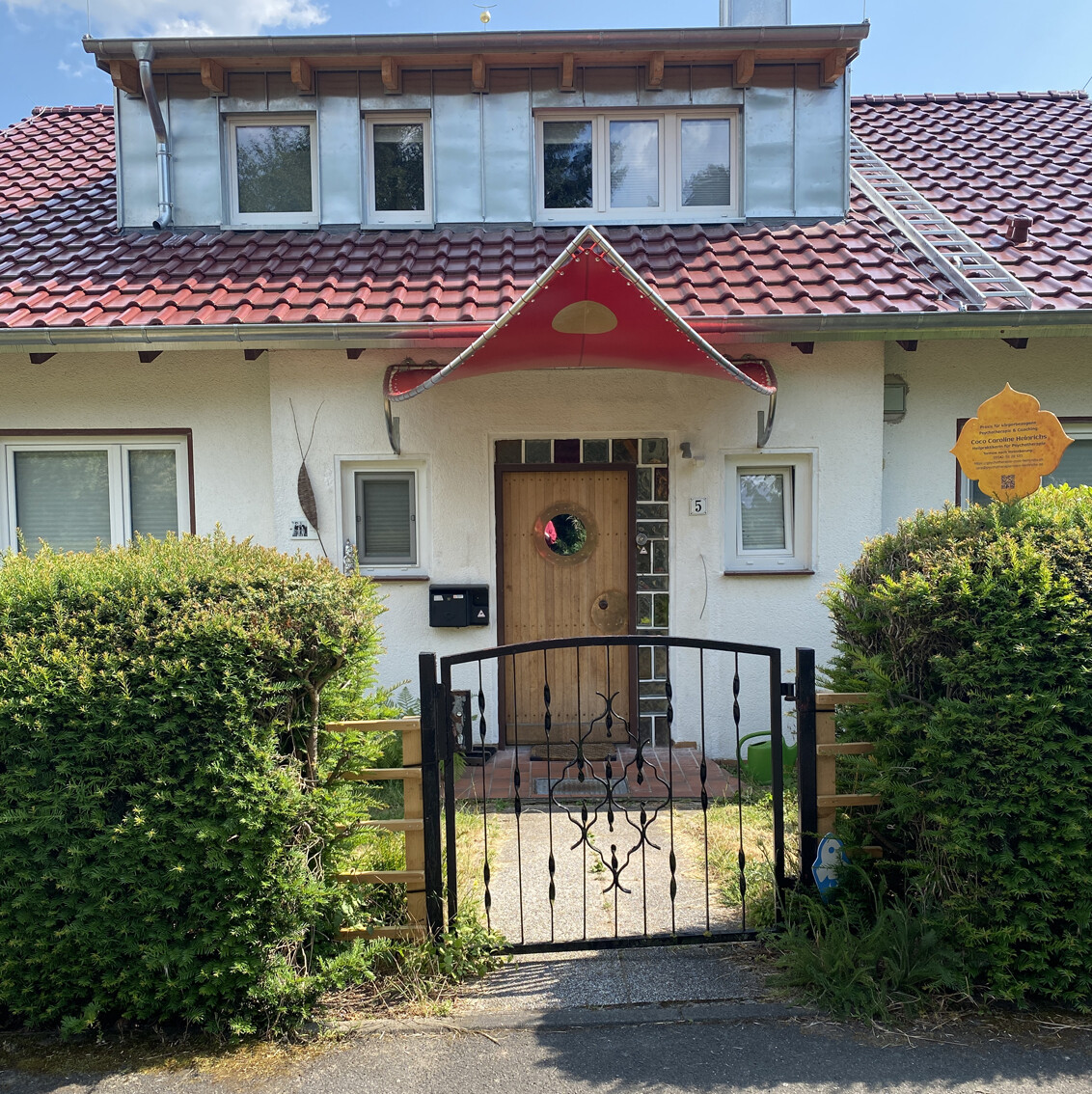 House front with red sail-like porch roof over wooden door, windows, garden fence, and hedges.