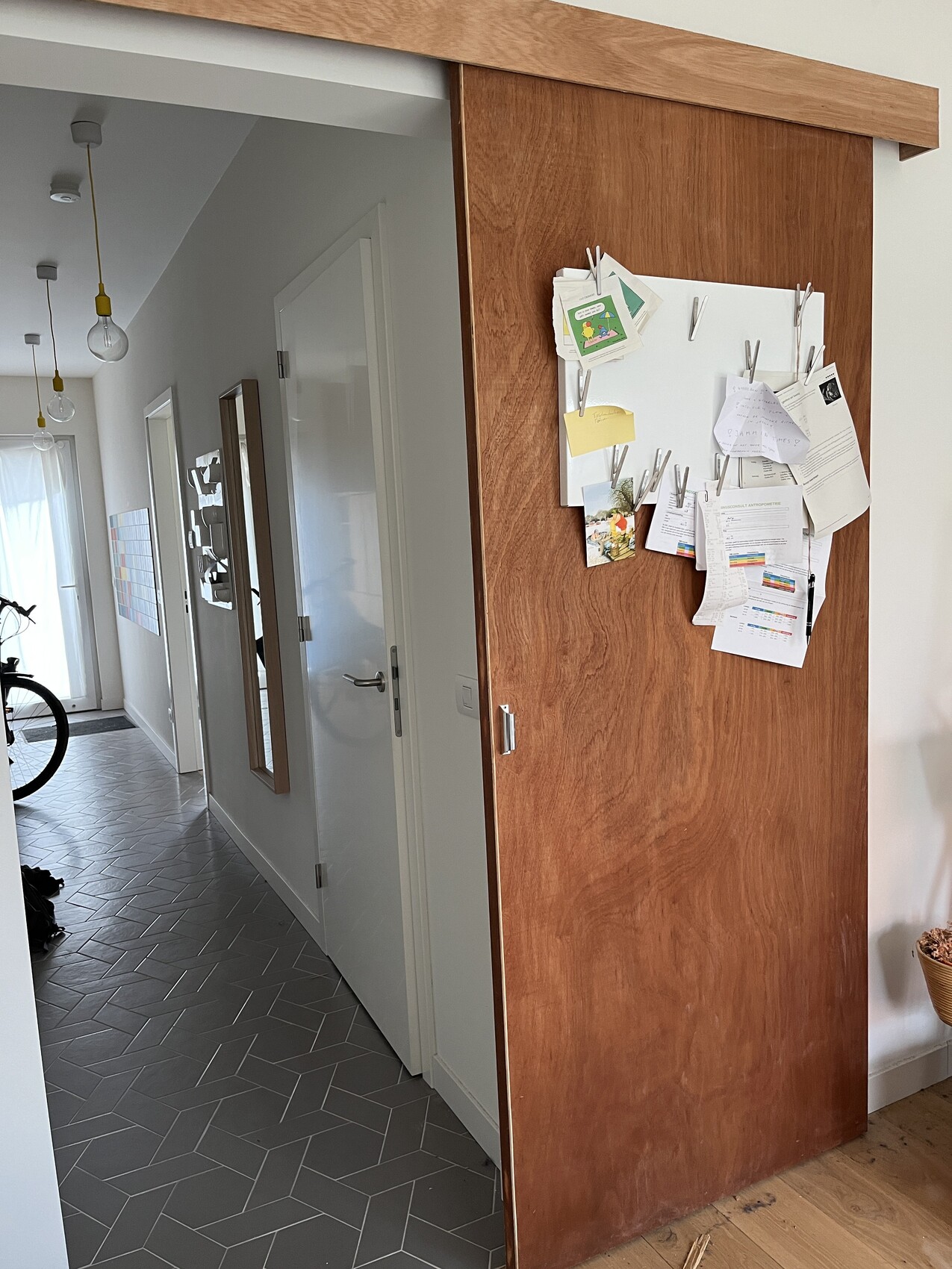 Bright hallway with gray tiled floor, wooden wall with a pinboard full of notes; bicycle on the left.