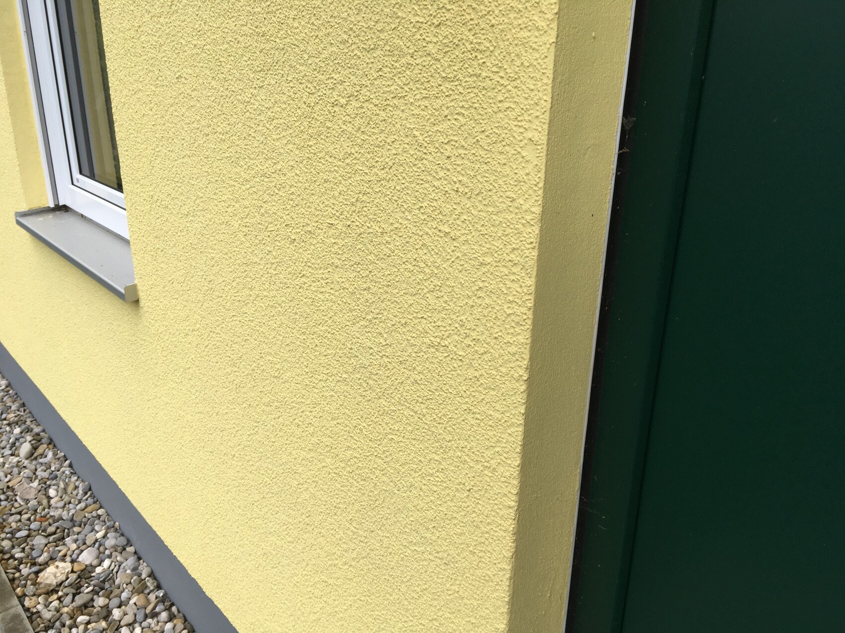 Exterior wall: yellow coarse plaster texture, window on the left, green door on the right, gravel ground.