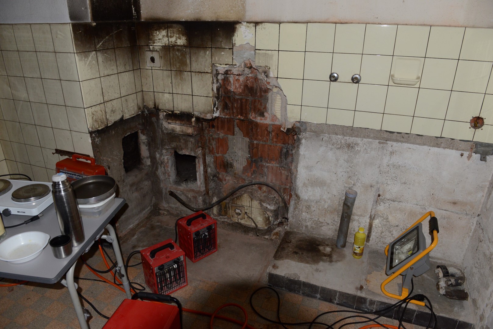 Kitchen area with damaged wall, tiles, pipes, and renovation tools.
