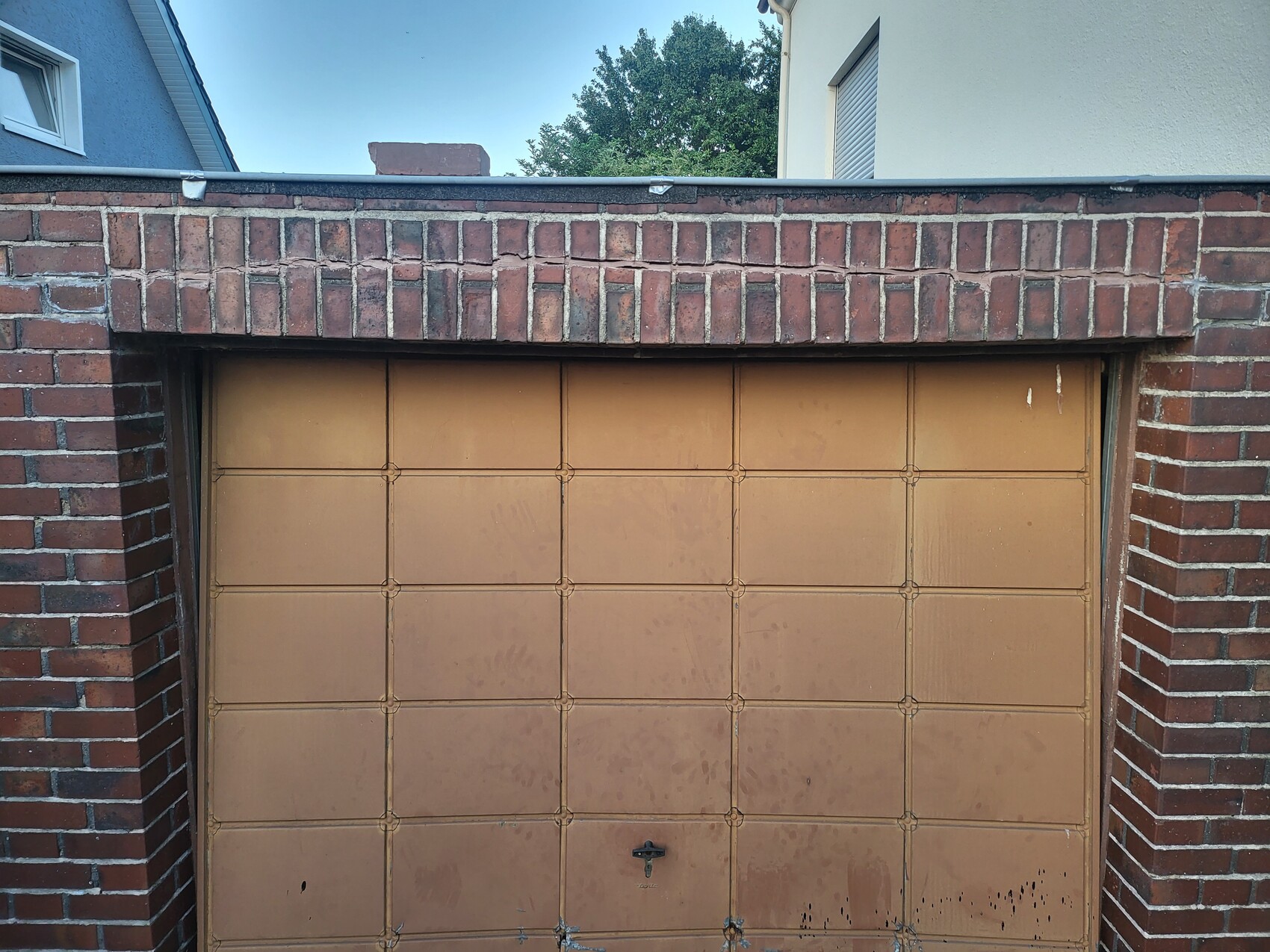 Brown, panel-style garage door in front of red brick wall; blue house on the left, light-colored building on the right.
