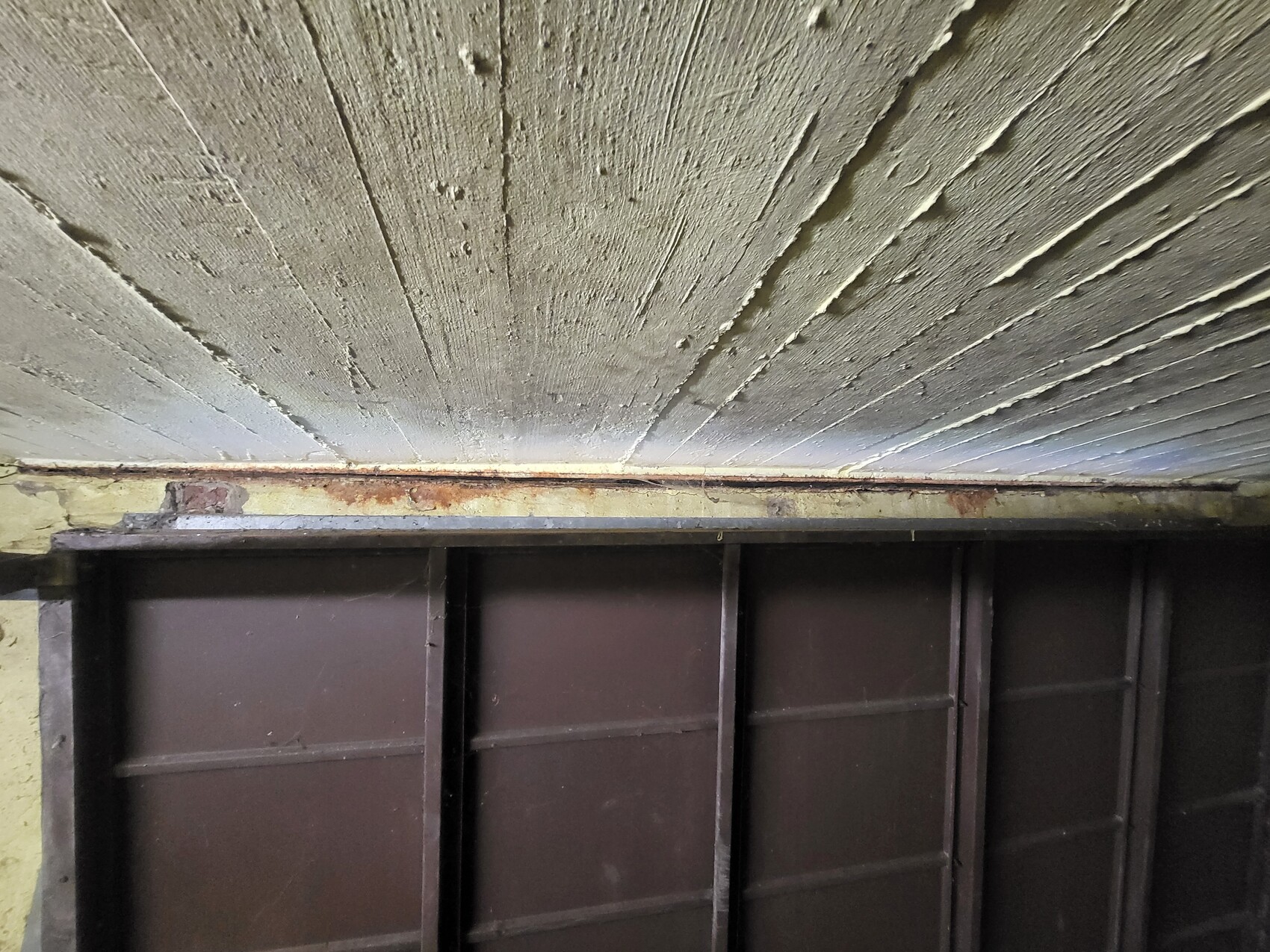 Underside of a roughly painted concrete ceiling with a rusty metal strip above brown wall panels.