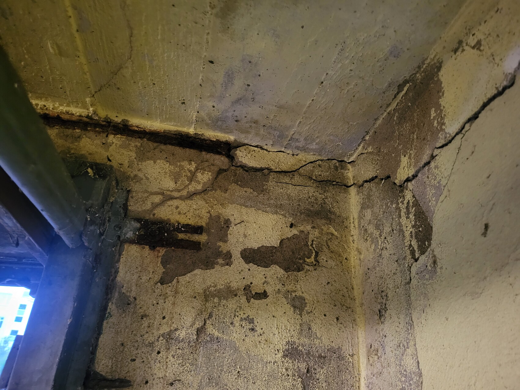 Basement corner wall made of concrete with cracks, moisture and stains; pipe on the left.