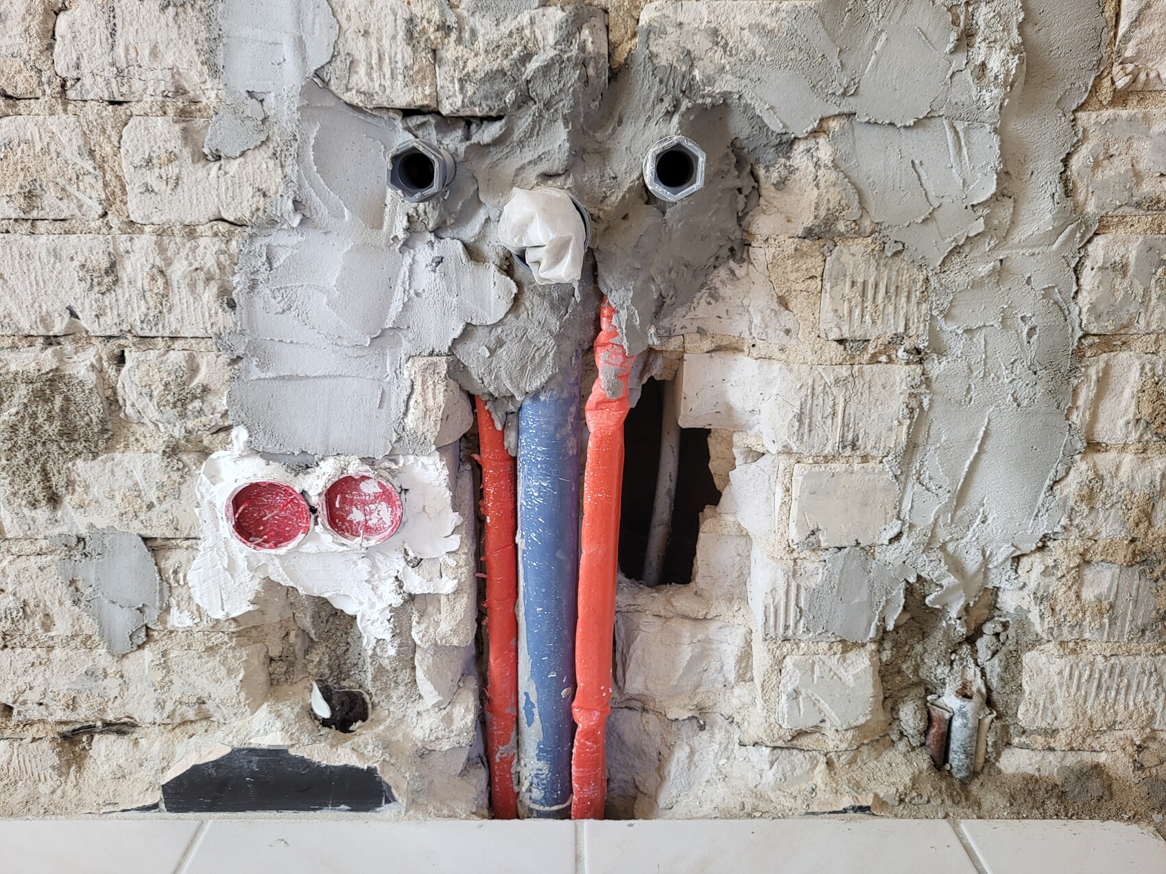 Open red and blue pipes in a damaged plaster wall with holes and bricks.