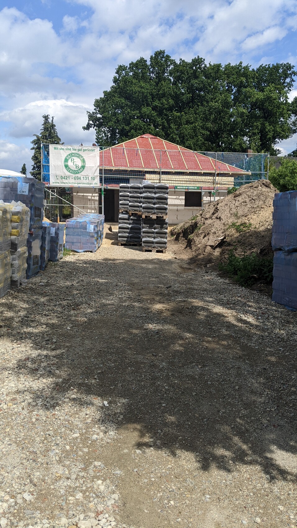 Construction site with stacked pallets, shell structure with red roof, dirt pile in the foreground.