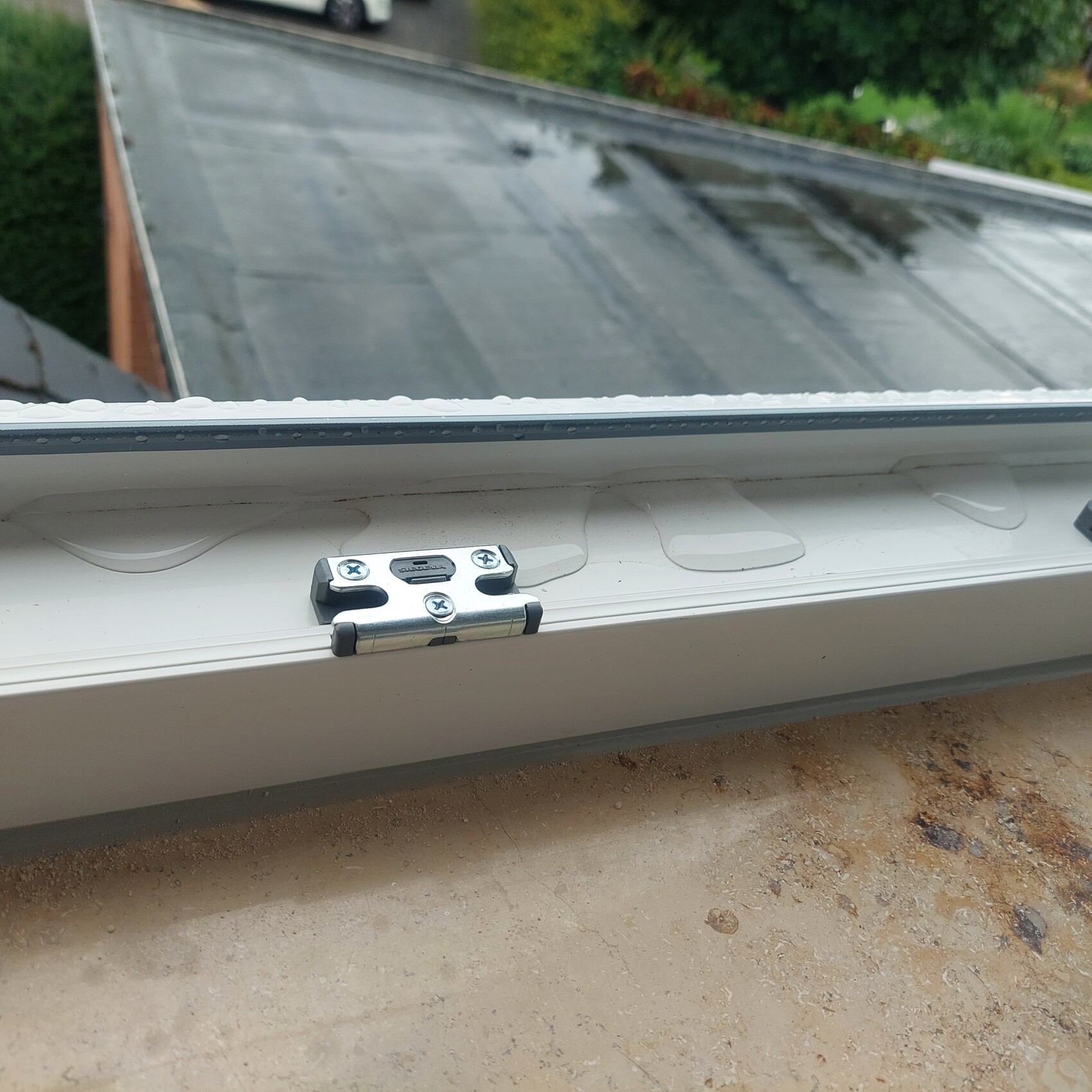 Roof window with silver metal hinge on white frame, rain-wet glass.