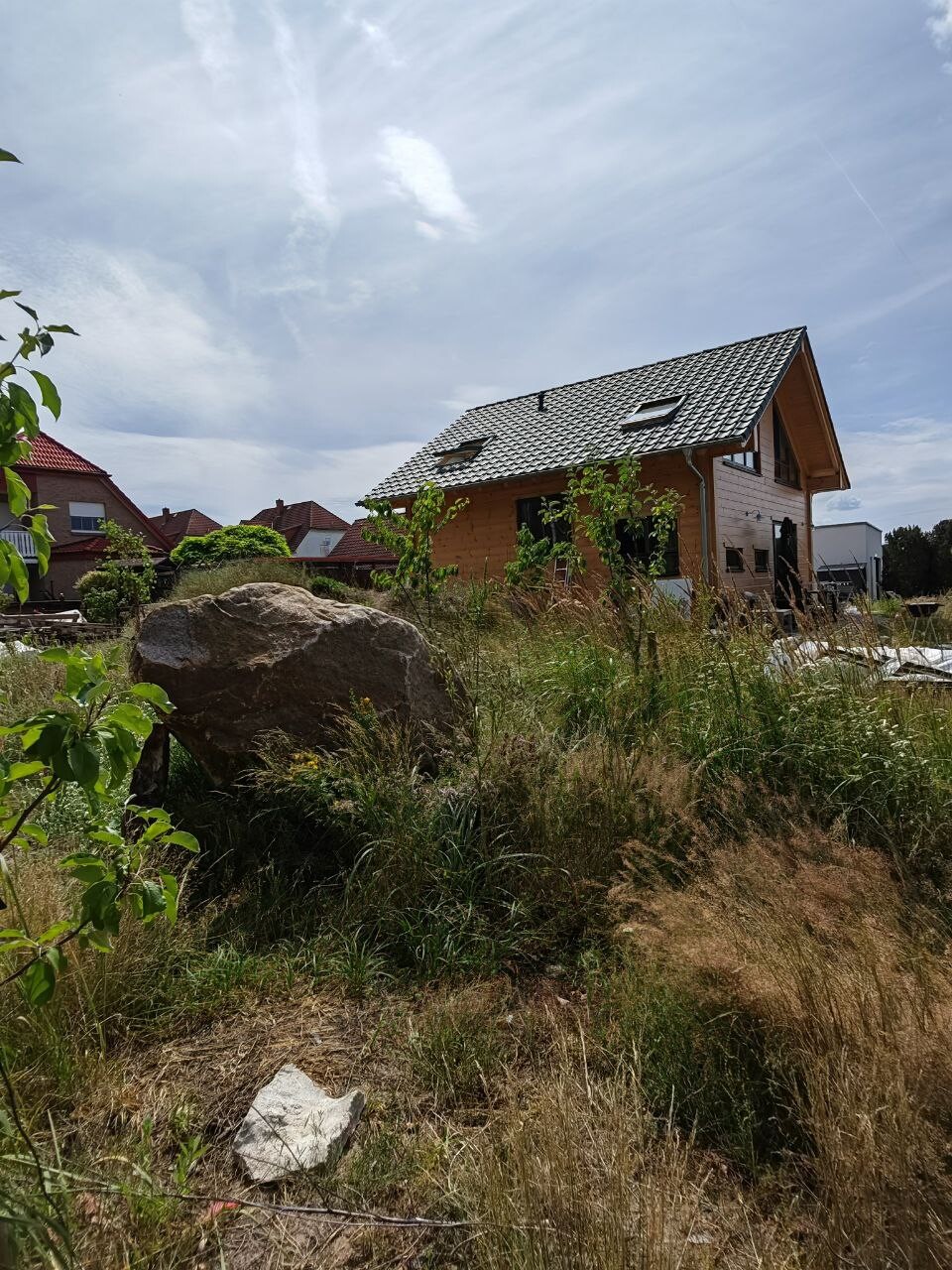 Wooden house under construction on grassy land, large boulder in the foreground, neighboring houses in the background.