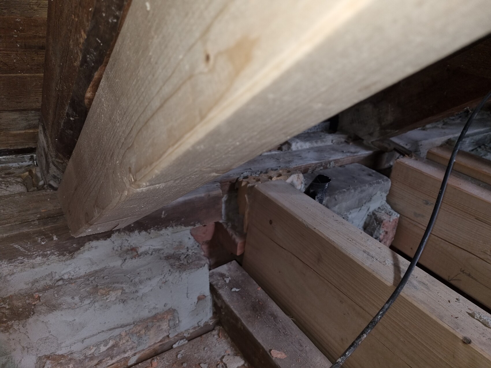 Attic with sloping wooden beam, wooden planks, brick fragments, dust and a cable.