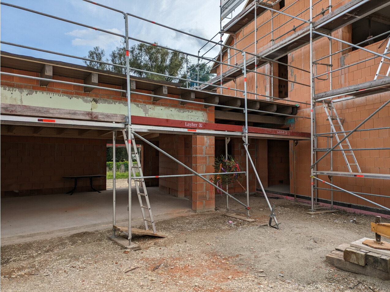 Shell construction made of red bricks with scaffolding and ladders; construction site with gravel floor and open door openings.