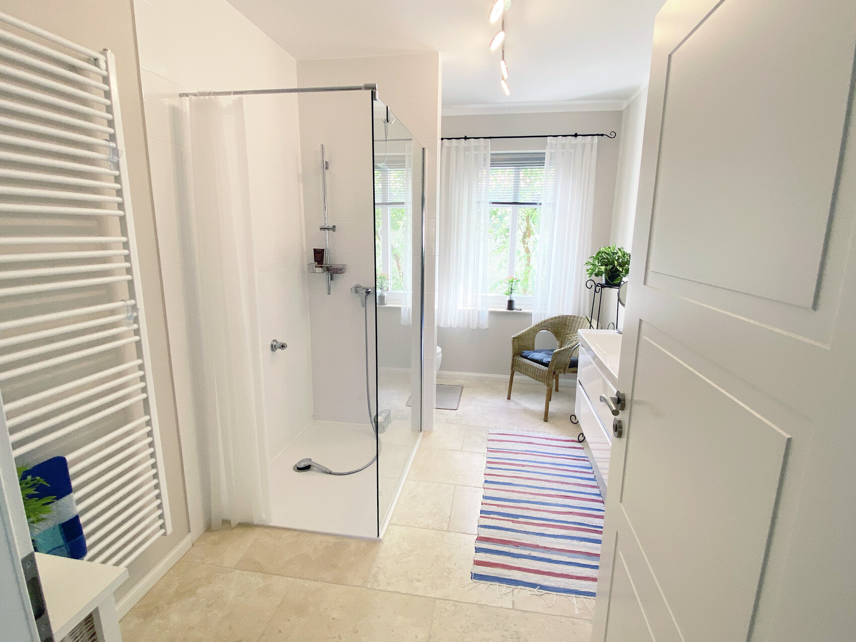 Bright bathroom with glass shower enclosure, window with curtains, wicker armchair, and striped rug.