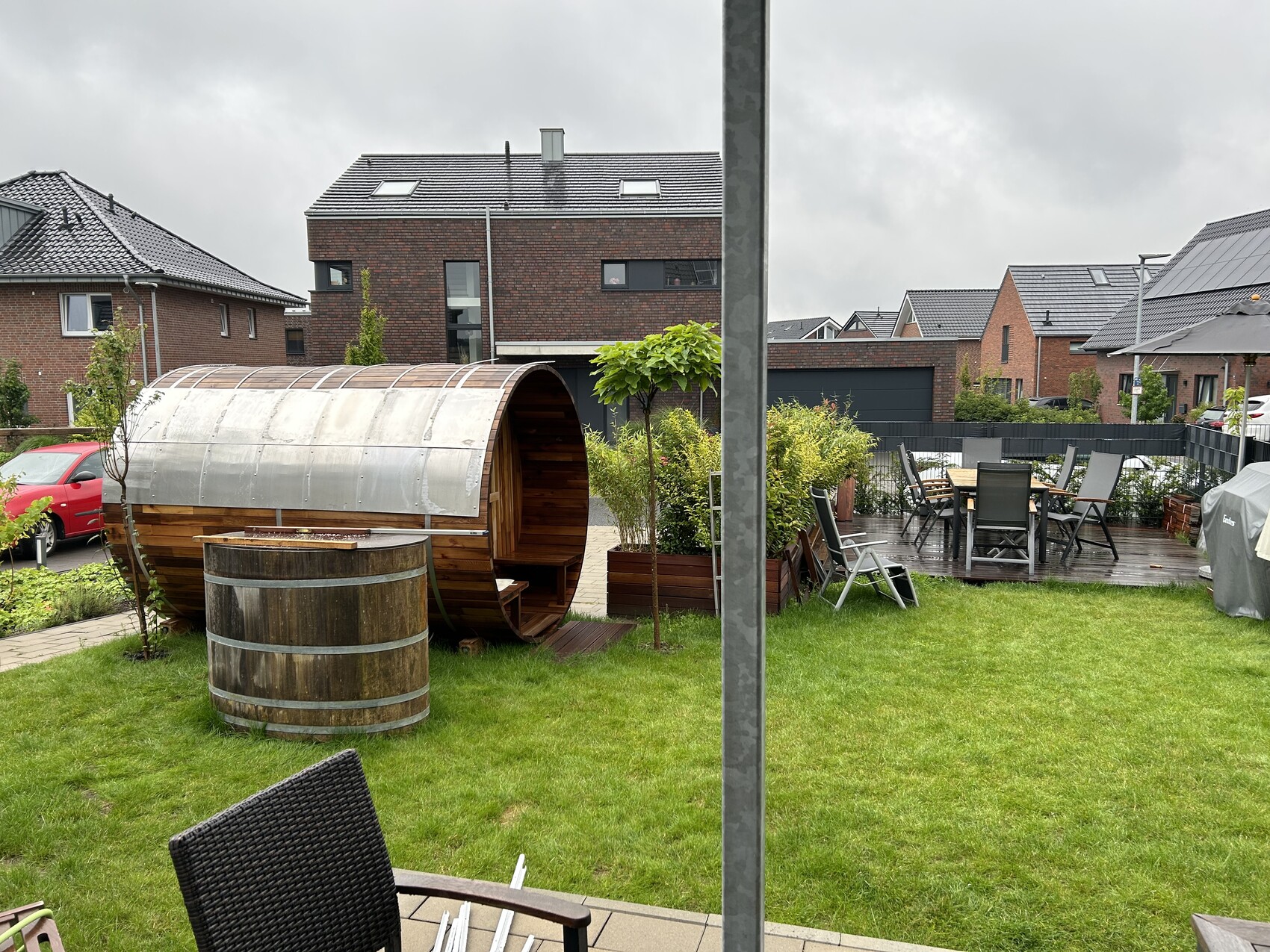 Garten mit Holzfass-Sauna, Esstischgruppe auf Terrasse, Liegestuhl; umliegende Wohnhäuser.