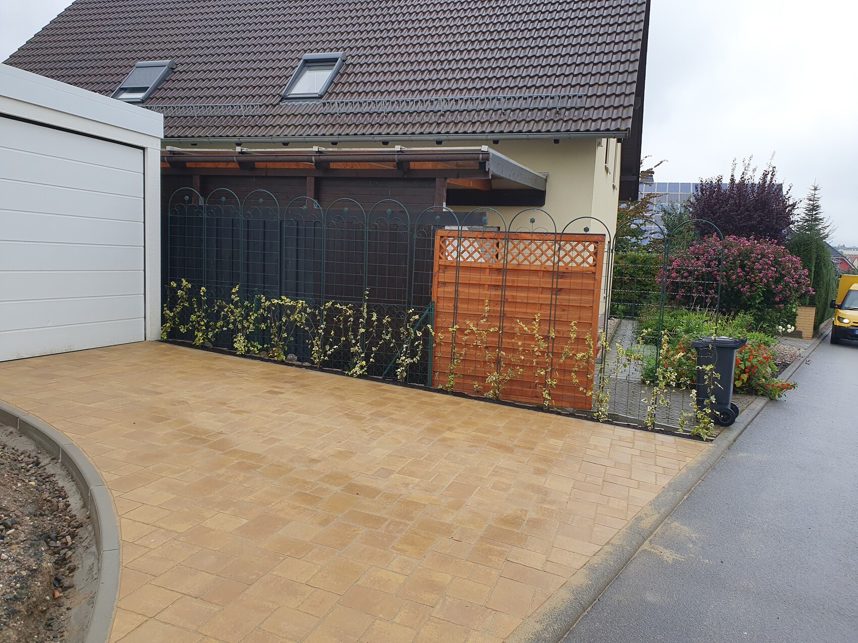 Front view of a paved driveway, garden fence, climbing plants, wood panel and trash bin.