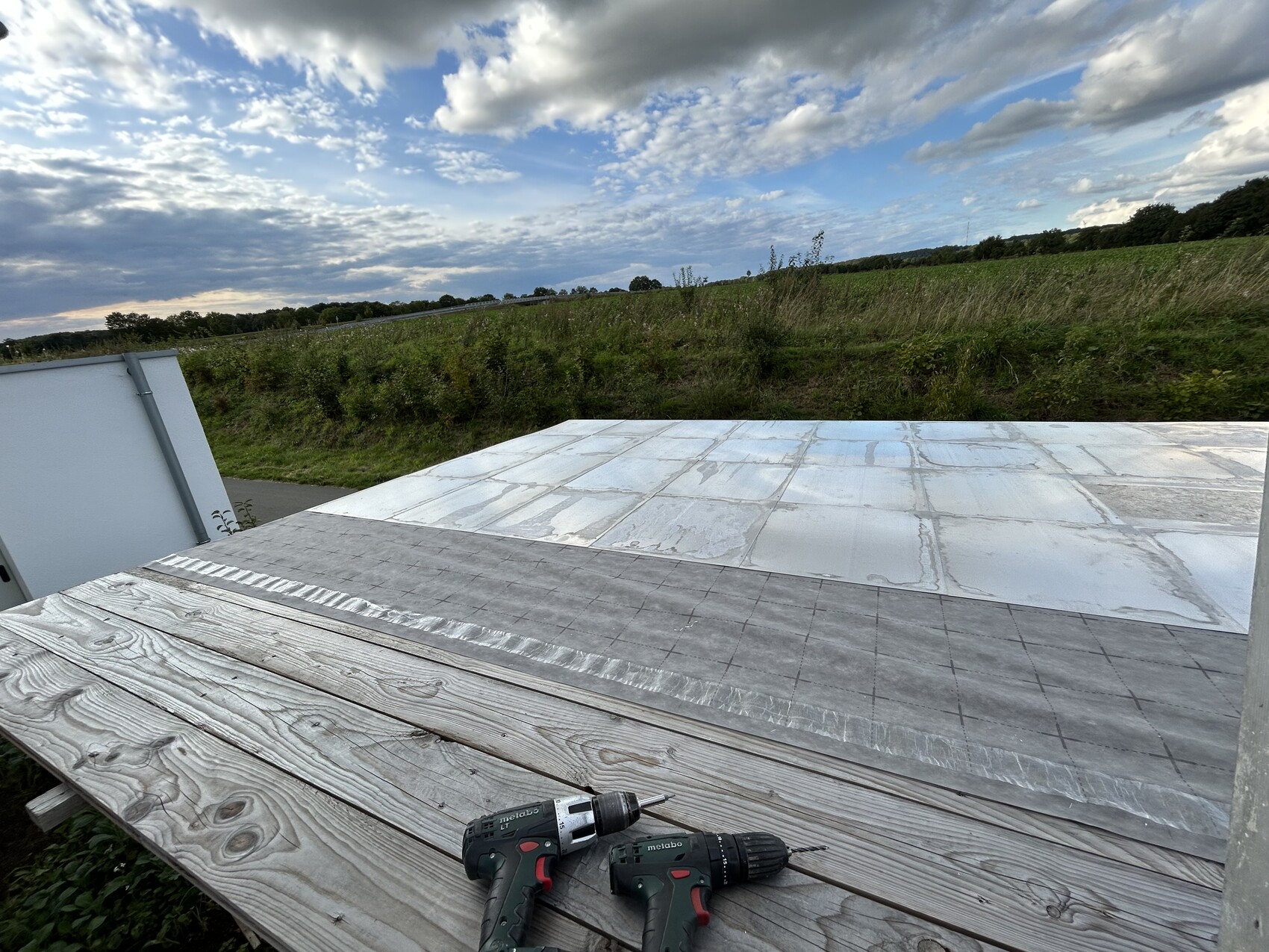 Wooden deck with two drills in front of a flat metal roof; fields and cloudy sky in the background.