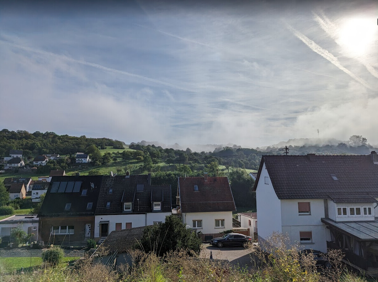View of houses on a slope, green fields behind, blue sky with contrails and sun.