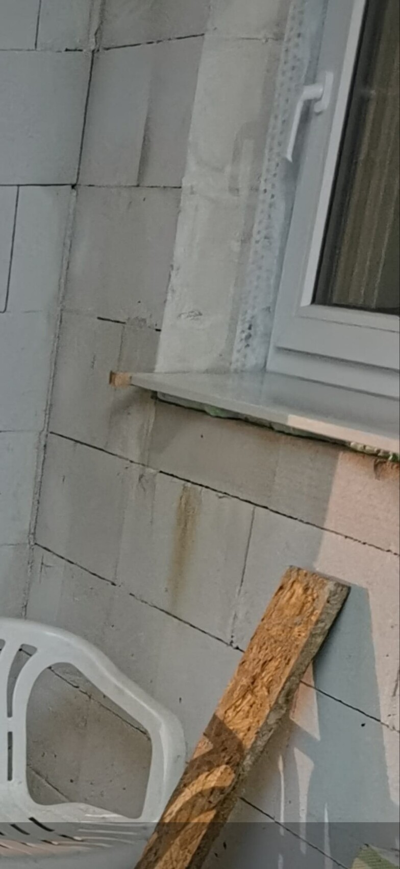 Exterior wall made of grey concrete blocks with window; wooden boards and plastic chair visible.