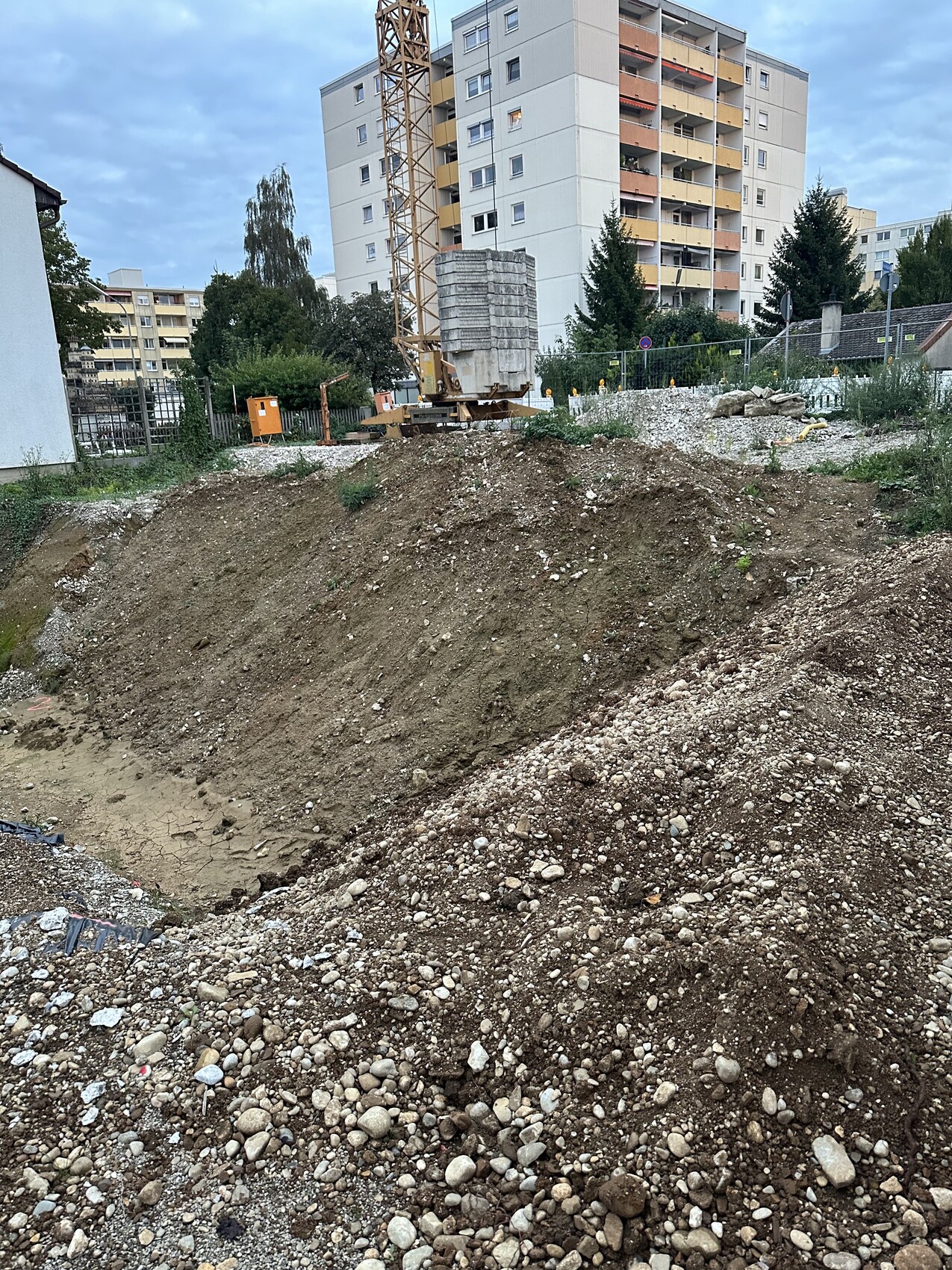 Construction site with soil piles, crane and concrete blocks, high-rise building in the background.